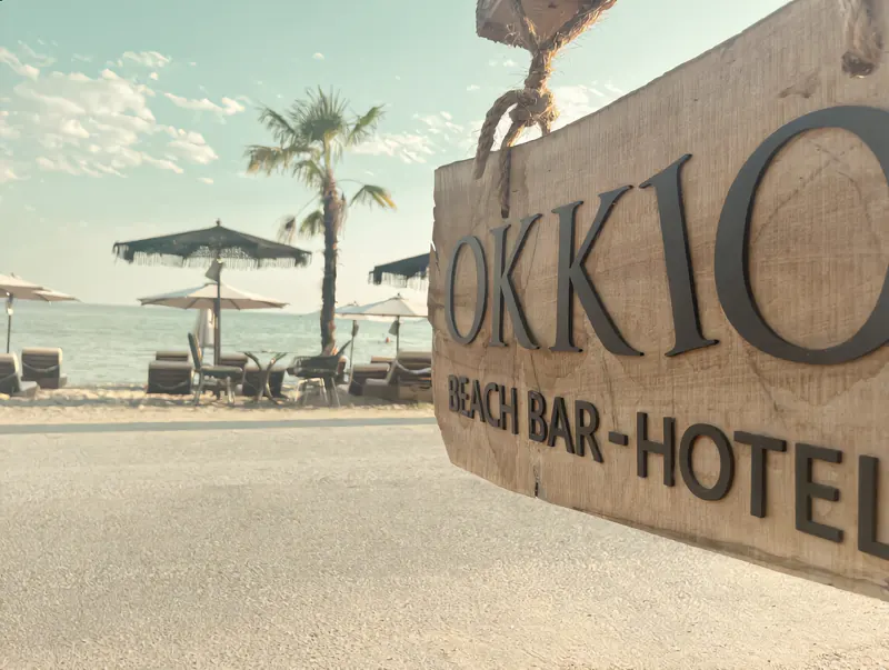 Wooden OKKIO Beach Bar sign with parasols and the sea behind